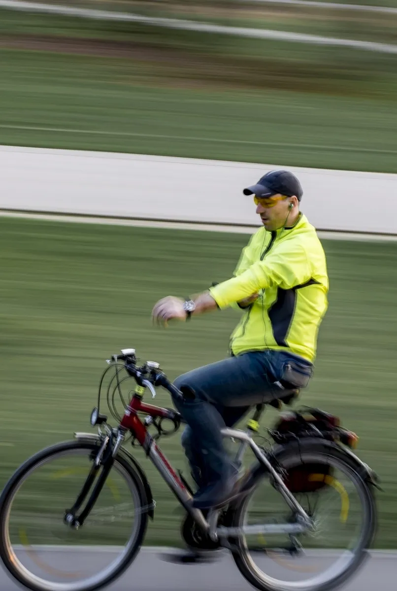 Cyclist in a neon jacket glancing down at an arm while riding through a blurred green background